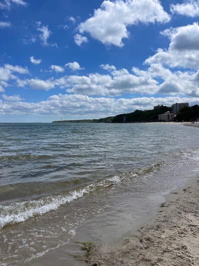 This media file captures a serene coastal landscape under a vibrant blue sky filled with numerous fluffy white cumulus clouds. The image is oriented vertically, showing the horizon line diagonally across the frame.
In the foreground and mid-ground, the calm, sparkling surface of a body of water (likely the Baltic Sea, given the location context) dominates, with gentle waves lapping at a sandy shore visible towards the bottom right. The water reflects the bright sky, creating shimmering patterns.
Along the distant horizon on the left side of the image, a lush, tree-covered hill rises, upon which several multi-story buildings are visible, suggesting a town or resort area. A faint, tall structure, possibly a tower or monument, can be seen on the distant water line in some crops.
The scene depicts a bright, clear daytime with pleasant weather conditions. The sandy beach and calm waters evoke a tranquil atmosphere. The setting is the coast of Pionersky, Russia. There are no clearly identifiable people or visible text within the image.
