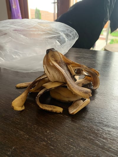 The image shows a heap of banana peels on a dark wooden surface, possibly a table. A crumpled, clear plastic bag is visible to the left of the banana peels. In the background, a window reveals a glimpse of the outdoors, possibly Kremyonki, Russia, suggesting a residential or indoor environment. No people are clearly visible in the frame, although a portion of a black garment suggests someone is present. There is no visible interaction or action.