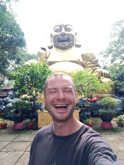 A man with a shaved head and a wide smile, eyes closed, takes a selfie in front of a large golden statue of the Laughing Buddha. The statue is seated and appears to be made of bronze. The man is wearing a dark grey crew neck t-shirt. Behind him, lush green foliage and potted plants surround the statue. In the background, a stone wall and a traditional Vietnamese-style wooden cabinet with ornate carvings are visible. The ground is paved with cracked stone tiles. The scene appears to be set in a temple or garden, likely in the city of Nam Ban, Vietnam.