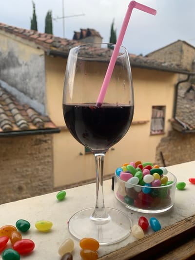 The image shows a glass of red wine with a pink straw, placed on a surface next to a small clear container filled with colorful jelly beans. Scattered around the container are several individual jelly beans in various colors like red, orange, yellow, green, and white. The glass of wine is partially filled, and the straw extends from it at an angle. In the background, there is a blurred view of buildings with tiled roofs, suggesting an outdoor setting. The sky is overcast, indicating a cloudy day. The overall scene appears to be a relaxed moment, possibly in a cafe or a residential balcony. The jelly beans add a playful and sweet element to the scene, contrasting with the sophisticated beverage. The location cues like the tiled roofs and architectural style of the buildings hint at a European setting, specifically San Quirico d'Orcia, Italy. There is no visible text in the image.