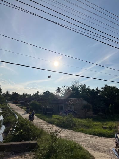The image depicts a rural scene in Alurcucur, Indonesia, under a bright, sunny sky with scattered clouds. Power lines are visible crisscrossing the upper portion of the frame. In the sky, a small object, possibly a kite or a distant drone, is flying. The landscape features a dirt road leading into a village, with several modest houses with tin roofs. Lush green vegetation, including palm trees, borders the road and surrounds the dwellings. On the road, a person is riding a motorbike away from the viewer. To the left of the road, a narrow body of water, likely a canal or irrigation ditch, is present. The overall impression is one of everyday life in a tropical village.