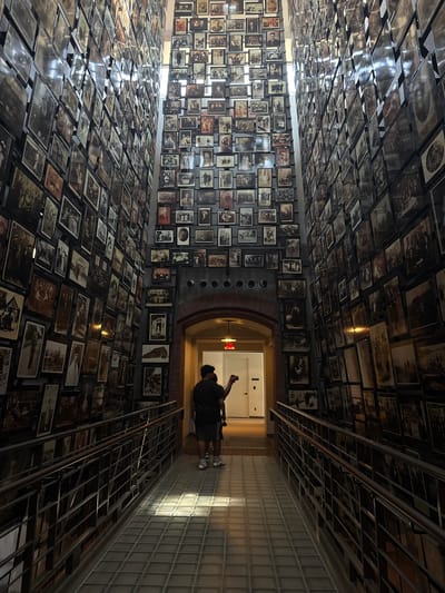The image depicts a museum exhibit with a vast collection of black and white photographs displayed on walls and an expansive floor installation. The photographs appear to be historical, showing people in various settings, possibly from the early to mid-20th century. The walls are covered with framed images, creating a mosaic effect, and the floor is covered with rows of unframed photographs, laid out on metal racks.
In the background, a person is visible walking away from the camera, towards a brightly lit hallway. This person is dressed in casual attire and appears to be a visitor to the exhibit. The lighting in the scene suggests it is daytime, with natural light filtering in from an unseen source, creating strong shadows on the floor and illuminating the glass block wall on the right. The overall atmosphere is somber and contemplative, fitting for a historical exhibition. There is no visible text in the image. The location is identified as Washington, United States of America.