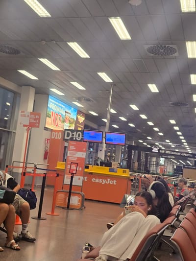 This is a photograph taken in the departure lounge of an airport in Somma Lombardo, Italy. The scene depicts a waiting area with rows of seating, illuminated by fluorescent ceiling lights. Several individuals are seated, some with headphones on, engaged with their phones or looking towards digital displays.
Prominently featured are several information screens and directional signage, including signs for "D10," "easyJet," and flight information indicating "Athens." One large digital billboard displays an advertisement with the text "PROVALA NO?".
In the foreground, a young woman is seated, leaning forward and looking at her mobile phone. She is wearing white pants and is partially obscured by her posture. Other people are visible in the background, some walking and others seated.
The environment suggests a typical airport departure lounge with barriers, check-in counters or gates, and advertising. The overall lighting is bright and functional.