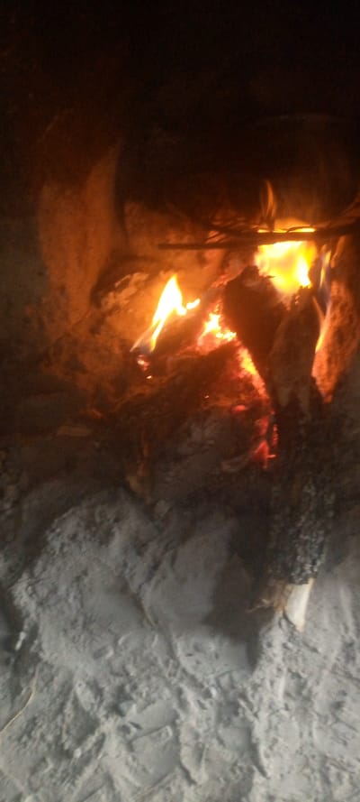 The image presents a close-up, warm-toned view of a fire burning in what appears to be a rustic fire pit or an enclosed fireplace. The scene is characterized by the intense glow of flames, contrasting with a dark background and a lighter foreground.
In the lower half of the frame, there is a substantial area of light grey, unevenly textured material. This appears to be a mix of fine ash, sand, or dirt, showing variations in shading and texture, with some areas appearing smoother and others more granulated, suggesting the ground of the fire-containing structure.
Above this ground layer, the mid-section is dominated by the actively burning fire. Several pieces of wood, appearing as logs or chunks, are visible amidst the flames and glowing embers. The fire exhibits a lively spectrum of colors, from deep reds and oranges in the smoldering parts and glowing coals to bright yellows and vivid oranges in the flickering, dancing flames that rise from the wood. The flames are dynamic and irregular, indicating active combustion, and they cast a strong, warm light onto the immediate surroundings.
The background, predominantly in the upper half of the image, is dark, ranging from deep browns to blacks. This area is subtly illuminated by the fire's intense glow, revealing indistinct textures that suggest the rough inner walls or enclosure of the fire pit or fireplace. There are no clear identifying features in the background beyond these shadowy, warmly lit contours.
The fire is the primary and most significant light source, creating a dramatic contrast between its bright, energetic warmth and the encompassing darkness. The overall impression conveyed is one of warmth, light, and perhaps a sense of primitive comfort in an otherwise dim or dark environment. There is no visible text in the image.