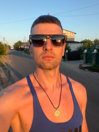 The image is a selfie of a man taken outdoors during what appears to be golden hour. He is wearing sunglasses and a blue tank top, with a necklace that has a circular pendant. The background shows a paved road, houses, trees, and a clear sky. The lighting suggests it is either sunrise or sunset. The location is identified as the city of Navahrudak, Belarus.