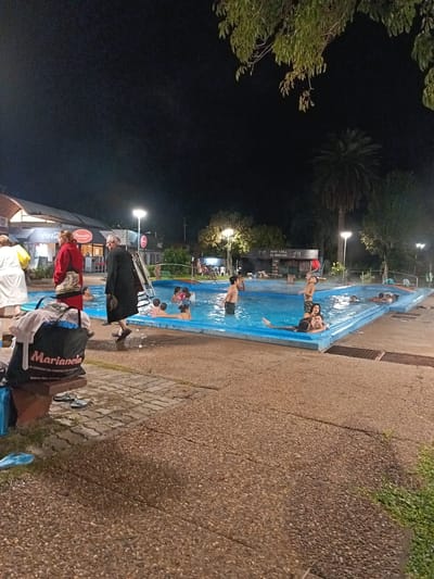 The image shows a nighttime view of a thermal pool in Termas del Daymán, Uruguay. People are swimming in the illuminated pool. Buildings and foliage surround the pool, with a "Coca-Cola Trouville" sign visible on a nearby structure. A person in dark clothing is walking near the pool edge. A bench with a bag labeled "Marianela" sits in the foreground. The ground is a paved surface. Overhead lighting illuminates the scene.