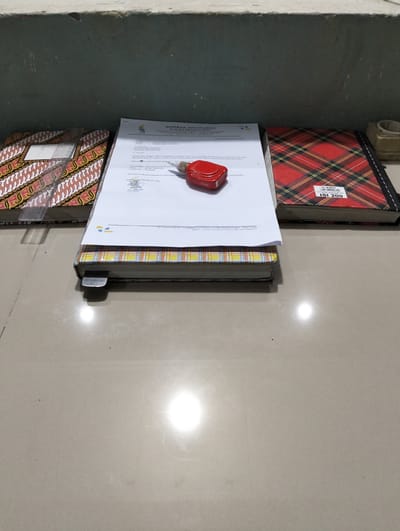 The image shows three stacked notebooks and a document on a floor. The top notebook has a red and black plaid pattern and a label that reads "C-KEY ISI 200". The middle notebook has a yellow and orange plaid pattern and a metal clasp. The bottom notebook has a batik pattern with red, yellow, and brown designs. A document with Indonesian text and a red stamp is placed on top of the middle notebook. The scene is lit by a bright overhead light source, casting reflections on the glossy floor. The background consists of a textured, dark wall. The location is identified as Tambontunong, Indonesia.