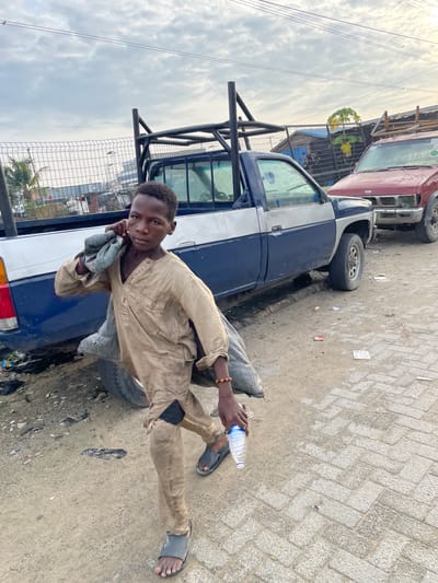 A young boy walks along a dirt and paved path in Ojomu, Nigeria, carrying a sack over his shoulder and a plastic water bottle in his hand. He is wearing a tan tunic and pants, and dark sandals. Behind him, a blue and white pickup truck is parked on the dirt shoulder, its bed equipped with a metal rack. Further back, a red pickup truck is partially visible. The environment appears to be an industrial or open area with a chain-link fence and some buildings in the background under a cloudy sky. Scattered debris is visible on the ground.