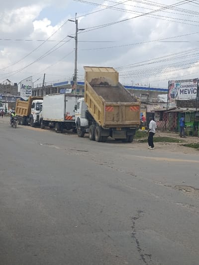 The image depicts an outdoor scene in Matungulu west ward, Kenya, under an overcast sky. An asphalt road, exhibiting cracks and patches, occupies the foreground and mid-ground. On the left side of the road, a line of four large commercial vehicles is oriented away from the viewer. These vehicles include, from left to right: a tan dump truck, partially obscured by a black motorcycle with a rider wearing a yellow-green reflective vest; a white box truck; and two white-cab dump trucks with raised yellow beds, each containing brown, granular material consistent with soil or sand.
On the right side of the road, a dirt shoulder borders the asphalt. A man in a white long-sleeved shirt and dark trousers stands on this dirt shoulder, facing right. Further right, a series of commercial structures are visible, including a small building with red and white checkered patterns, near which a person in a red shirt is present. Another individual in a blue shirt stands near a green structure further down the road.
In the background, a large blue and white building is partially visible. A tall sign for "JEXO" stands on the left, and a large billboard displaying "CAMBRIDGE" and "MEVIC" is prominent on the right. Multiple utility poles support numerous overhead power lines that traverse the scene.