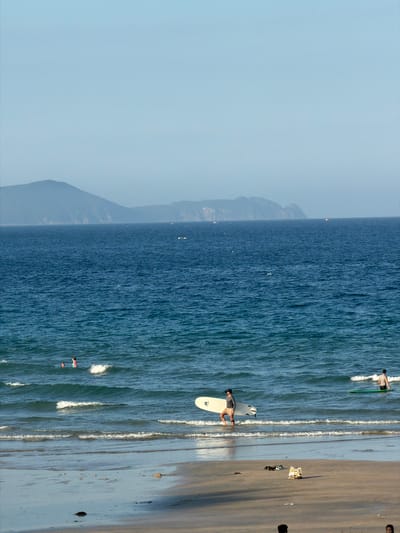 The image depicts a coastal scene in Cam Lam, Vietnam, likely on a bright, clear day. The foreground is a sandy beach with a few scattered objects, possibly debris or personal belongings. The water is a vibrant blue, with gentle waves lapping at the shore.
In the water, several individuals are visible. To the right, a person is standing in the water with a surfboard. Further to the left, a person is walking out of the water carrying a surfboard. Two other people are in the shallow water to the left, one appearing to be a child in a pink swimsuit.
In the distance, the expansive blue ocean stretches to the horizon, where a hazy line of mountains forms the backdrop. The sky is a pale, clear blue, with no clouds visible. The overall impression is one of a serene beach day.