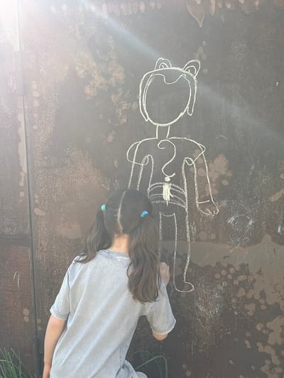 In Novaya Usman, Russia, a young girl with dark brown hair styled in two pigtails, adorned with blue hair ties, is seen from behind. She is wearing a light gray t-shirt and is actively drawing a chalk outline of a figure on a large, weathered, rusty metal surface, possibly a wall or a door. The chalk drawing depicts a stylized human figure with large ears, a question mark on its chest, and a belt. Sunlight streams down from the upper left, creating a lens flare effect and illuminating the scene. The background is a rough, textured surface with visible rust stains and a vertical seam, indicating an outdoor industrial or weathered urban setting. Some sparse green vegetation is visible at the bottom left of the frame.
