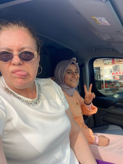 Two women are in a car, likely in Şile, Turkey. One woman is taking a selfie while sticking her tongue out and wearing sunglasses and a white shirt with embellishments around the neckline. The other woman is lying in the back seat, wearing a hijab and an orange sweater, smiling, and giving a peace sign. Through the car window, a building with a sign that says "KASA" in large letters is visible.