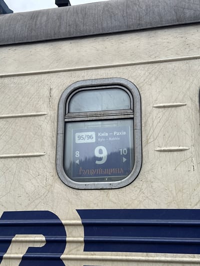 A close-up, slightly rotated image captures the weathered exterior of a train car, likely in Tatariv, Ukraine. The main body of the car is a light beige or off-white, covered in numerous scratches and marks, suggesting considerable use and age. To the right, a vertical dark blue stripe runs down the train's side, partially revealing a large white letter, possibly "B" or "Я".
The central feature is a rectangular display window set into the train's side, framed by a worn metallic border. Inside, against a blue-grey background, white text provides crucial information. The route is indicated as "Київ — Рахів" (Kyiv — Rakhiv) in both Ukrainian and English. Below this, the train number "95/96" is visible. A large, prominent white number "9" identifies the train car number, accompanied by smaller numbers "8" and "10" with directional arrows, likely indicating adjacent car numbers. Vertically displayed, the name "ГУЦУЛЬЩИНА" (Hutsulshchyna) identifies the train, referencing the historical Hutsul region.
The overall lighting is even, suggesting an overcast daytime sky. The image focuses on the functional signage of a train, providing clear identifying details for its route and car designation. No people are visible, emphasizing the train's structure and informational panel.