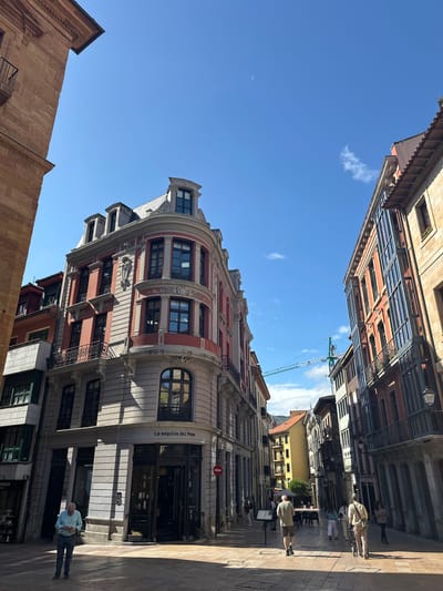 This is a photograph of a street scene in Oviedo/uviéu, Spain, taken on a clear, sunny day. The low angle of the shot emphasizes the architecture of the buildings lining the street, which are predominantly old and ornate, with a mix of stone and stucco facades. Some buildings feature balconies with decorative ironwork, and the windows are tall and multi-paned.

In the foreground, a man in a light blue shirt and dark pants stands on the sidewalk, appearing to be looking at his phone. The street is paved with cobblestones, and the sunlight casts long shadows, indicating it is likely late afternoon.

Further down the street, other people can be seen walking and standing. The buildings on either side of the street create a narrow canyon effect, with the blue sky visible above. One building in the midground is a vibrant red and cream color, with a curved facade. There is a sign on this building that reads "La esquina del Peso."

In the background, a construction crane is visible against the bright blue sky, suggesting ongoing development in the area. The overall atmosphere is one of a typical European city street, with a blend of historical architecture and modern life.