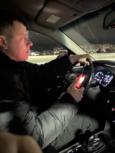 A person is inside a car, looking up and towards the left. They are wearing a black jacket and a light grey puffer jacket over it, as well as a watch. The person is holding a phone with a red case in their right hand on the steering wheel. The car's dashboard and a night cityscape are visible through the windshield.