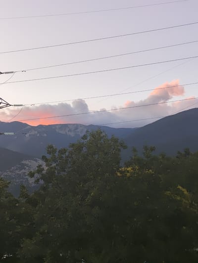 This image captures a serene mountain landscape at dusk, with power lines crisscrossing the frame against a soft, gradient sky of pale violet and pink hues. In the distance, several layers of mountains are visible, their slopes receding into the twilight. A band of warm, orange-pink light emanates from behind the peaks, suggesting the last rays of the setting sun. The foreground is dominated by the dark silhouettes of dense trees and foliage, with hints of lighter green and yellow interspersed among the leaves, possibly indicating flowers or the changing colors of autumn. The overall scene is calm and natural, with the man-made element of the power lines providing a subtle contrast to the rugged beauty of the mountains. The location is specified as Massandrovsky settlement council, Ukraine.