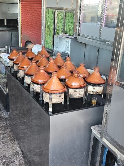 This image depicts an outdoor or semi-open commercial food preparation area in Boufakrane, Morocco. In the foreground, thirteen traditional earthenware tagines with conical lids are arranged on a long, reflective black counter. Each tagine rests on an individual, blackened metal charcoal burner, suggesting active cooking. A small glass containing an amber liquid is positioned on the counter near the rightmost tagine. To the left of the tagines, a glass display case is visible. In the background, a male individual, wearing a light-colored garment, leans over a stainless steel counter, engaged in an unseen task. Behind him, a partially open red metallic roll-down shutter reveals additional kitchen equipment. To the right of the individual, several windows with a green, reedy design are visible, along with a reflective grey cabinet or service window. The floor visible in the lower left consists of dark, patterned tiles.
