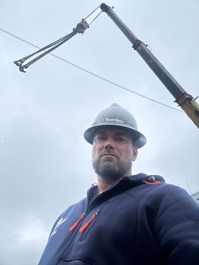 The media file is a selfie of a man with a beard and a hard hat, taken from a low angle looking up at the sky. He is wearing a dark blue hooded jacket with orange zipper pulls. The hard hat has a striped pattern and the text "Y.V. Bostrikov" is visible on it. To the left of the man, a crane boom extends upwards, with chains and hooks hanging from it. The background is a pale, overcast sky, with a faint outline of a building or structure visible at the top right. The overall lighting suggests it is daytime, possibly late morning or afternoon, with cloudy weather. The man's expression is neutral. The image appears to have been taken outdoors, likely at a construction site or industrial area, given the presence of the crane and hard hat. The location context provided is Usinsk, Russia.