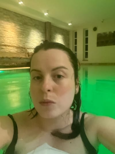 A woman with dark, wet hair is angled in the foreground, wearing a black swimsuit with a white towel. She is looking directly at the camera with a slight pout. Behind her is a green-tinted pool and a wall with multiple rows of windows, some of which have black vents. The room has recessed lighting in the ceiling. The wall's surface appears to be made of stone or brick.