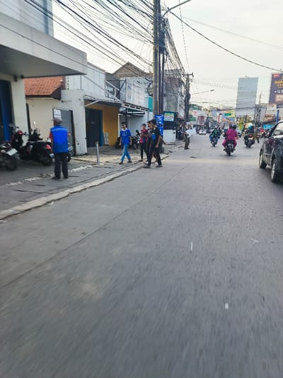 The image shows a street scene in Bojongsoang, Indonesia, captured from a moving vehicle. It is a sunny afternoon, with a clear sky and minimal cloud cover. There are several buildings lining the street, including a single-story building on the left with a blue facade and a sign hanging above the entrance. The buildings are mostly residential or commercial.
There are several people walking along the sidewalk on the left side of the road. They appear to be interacting with each other. There are also motorcycles parked along the side of the road, and several motorcycles are moving along the road in the distance. The road is paved with gray asphalt. There are numerous electrical wires overhead.
The image was captured from the perspective of a vehicle moving along the road, giving a view of the street and the surrounding buildings. The focus is on the scene in front of the vehicle, which is partially blurred due to the movement of the vehicle. The background is more distant and less clear. The buildings and other objects in the distance are visible, but they are not as sharp as those in the foreground. The image gives a glimpse of daily life in Bojongsoang, Indonesia.