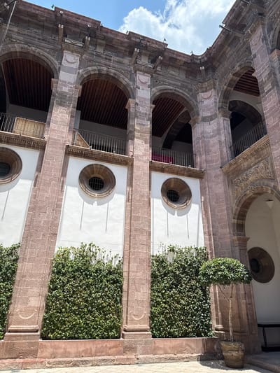 Here is a detailed description of the image:
The image depicts a vertical, upward-angled view of a multi-story building facade in San Miguel de Allende, Mexico. The building is constructed with a mix of light-colored stone, likely sandstone, and white-painted walls. The architectural style includes arched openings, some with railings, revealing interior spaces with wood beams. There are round windows with grilles set into the white wall sections. Planter boxes covered in lush green foliage vertically line the building's exterior. The lighting suggests it is daytime with clear shadows. The sky is partially visible in the upper left corner with a few clouds. There is no visible people in the image.