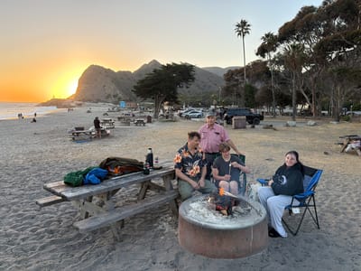 The image depicts a sunset beach scene at Leo Carrillo State Park in Malibu, California. In the foreground, four people are gathered around a fire pit on the sand. An older man in a pink button-down shirt stands behind a younger man and a woman. The younger man, wearing a dark t-shirt, is holding a stick with a marshmallow over the fire. The woman, seated in a blue camping chair, is wearing a black hoodie and grey sweatpants and smiling at the camera. To the left, another man in a floral shirt and green pants is sitting on a picnic table, next to a cooler and some blankets. The picnic table is made of weathered wood. In the background, the ocean waves are visible, and a rocky hill with trees and a road on it rises behind the beach. Several palm trees and other vegetation are scattered throughout the scene, and cars and more picnic tables are present in the distance. The sun is setting, casting a warm orange glow over the landscape.
