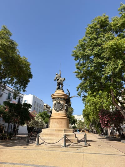 This is a low-angle, full-shot, exterior photograph of a monument in a public square in Eivissa, Spain. The monument is made of bronze and stone, depicting a figure on horseback wielding a sword. The monument is situated in the center of the frame, with a paved walkway extending outwards. Two large, leafy green trees frame the monument, one on each side. In the background, there are several white, multi-story buildings with balconies, typical of the architecture in Eivissa. The sky is a clear, bright blue. There are a few people scattered throughout the square, some sitting at tables outside, others walking. The scene is illuminated by bright sunlight, casting shadows on the ground. The overall impression is one of a well-maintained public space on a sunny day.