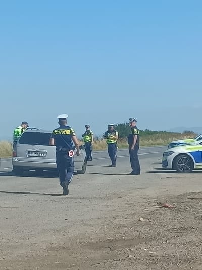 The image depicts a roadside scene in Tenevo, Bulgaria, under a clear blue sky. Several police officers in uniform are visible, some directing traffic or observing vehicles. A silver minivan is stopped on the asphalt road, with at least two officers interacting near it. Another police vehicle, a white and blue car with police markings, is parked further down the road. The surrounding environment appears to be rural, with dry grass and distant hills visible. One officer in the foreground is walking away from the camera, wearing a blue shirt with "POLICE" lettering and a white hat, and holding a red and white traffic control device.