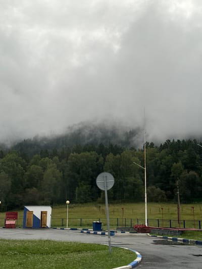 In Катунь, Russia, on an overcast day, a grey sky looms over a dense forest of green trees. The forest is partially obscured by fog, creating a misty atmosphere. In the foreground, a paved area with white and blue painted curbs leads to a small, blue and white building with two wooden doors. A red metal structure is located to the left of the building, and a blue trash receptacle is to the right. A tall, thin metal pole with a pointed top stands in front of the forest, accompanied by a street lamp. A circular sign on a pole is positioned in the middle-left of the frame. Beyond the building and poles is a grassy field, bordered by a fence with red piping.