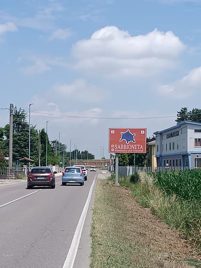 The photo shows a street in Mezzana Sant'Antonio, Italy, with a sign indicating the entrance to Sabbioneta, a UNESCO World Heritage Site. The sky is blue with scattered white clouds. A brown hatchback, a light blue subcompact car, and a white sedan are traveling along the asphalt road. A grassy verge is on the right side of the road, and a building is visible behind the sign. The street is lined with utility poles and streetlights.