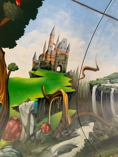 This is a close-up, distorted, overhead photograph of a mural painted on a glass surface. The mural depicts a fantastical, dreamlike landscape. Dominating the left side is a multi-turreted, tan and grey castle with pointed spires and square windows, seemingly built from blocks or dice. It sits on a green, undulating hill. To the right of the castle, a large, vibrant green, organic shape curves upwards, resembling a stylized plant or mountain. Further to the right, a waterfall cascades down a rocky cliff face into a dark, reflective pool. Other elements of the surreal landscape include abstract, wood-like structures, large, spotted red mushrooms, and rock formations. A dark green, jagged-topped tree with a red orb resembling a fruit or planet is visible in the lower left. The sky is painted with soft blues and whites, suggesting a daytime scene. The mural appears to be viewed through a clear, possibly reflective material, with horizontal lines, possibly the edges of glass panels or wires, bisecting the image. The overall impression is one of an otherworldly, imaginative realm. The location context is Odivelas, Portugal.