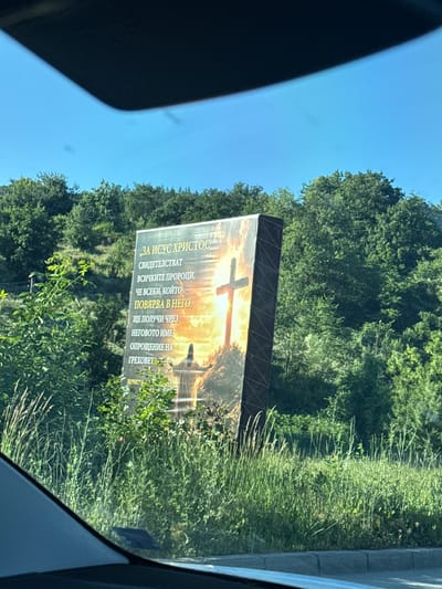 The image displays a large billboard featuring a religious message and imagery. The billboard is situated outdoors, with a backdrop of lush green trees and a bright sky. In the foreground, tall grass and foliage obscure the lower portion of the billboard. The billboard itself is tilted and shows a depiction of a cross with a silhouette of a person standing before it. The text on the billboard is in Bulgarian and appears to be a religious quote or scripture. The location is identified as Veselinovo, Bulgaria. The scene is captured from a vehicle, as indicated by the car's windshield and dashboard in the lower part of the frame.