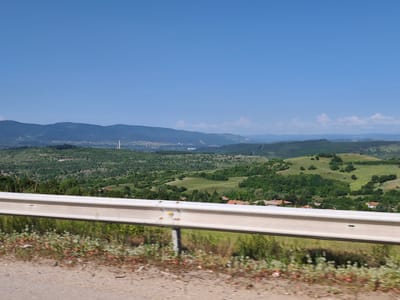 The image depicts a scenic view from a roadside guardrail overlooking a valley with rolling hills and a distant city skyline. The foreground features a light-colored metal guardrail, its surface showing some wear and tear, with sparse vegetation including dry grass and small reddish-green plants growing along the edge of the dirt road.
The middle ground reveals a lush green landscape with trees and scattered houses with red tile roofs, characteristic of a rural or semi-rural setting. The hills are covered in dense vegetation, with some patches of lighter green indicating open fields or meadows.
In the far distance, a hazy cityscape is visible, with buildings and infrastructure stretching across the horizon. A prominent, tall, slender structure, possibly a tower or chimney, stands out within the urban area. Mountains form the backdrop, fading into the blue sky. The sky itself is a clear, vibrant blue with a few wispy white clouds scattered towards the horizon.
The image's perspective suggests it was taken from a moving vehicle, as evidenced by the slight motion blur in the foreground vegetation. The overall impression is one of a sunny day, offering a panoramic vista of the Bulgarian countryside near Golemo Buchino.
