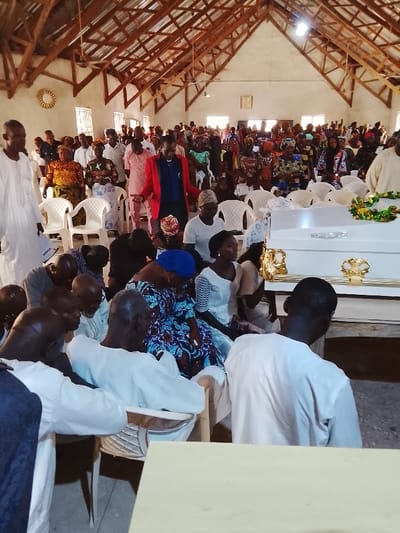 The image shows a funeral ceremony taking place inside a building with a wooden roof. It is likely the building is a church or community hall. The mourners are sitting in rows of white chairs in the foreground, some are praying or consoling each other, while others are seated in quiet contemplation. A white coffin with gold trim is positioned near the center of the image. There are floral arrangements near the coffin. The mourners are dressed in a variety of traditional African clothing, including colorful dresses and headscarves. The overall mood is solemn and somber, reflecting the nature of the event. The location is not Mukaiya, Nigeria. Mukaiya is a village and is not located in a city. There are no other identifying details in the image or the provided location coordinates to suggest the location of the ceremony.