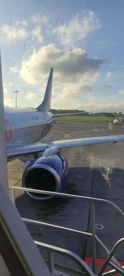 The image shows the side and engine of a commercial airplane parked on the tarmac at Khrabrovo Airport in Khrabrovo, Russia. The aircraft's tail fin, bearing the registration "RA-73555," is visible. The engine, painted in blue and silver, is prominently featured in the foreground. In the background, other aircraft, including a small helicopter, are visible on the runway. The sky is partly cloudy with a mix of blue sky and white cumulus clouds. The airport environment is characterized by asphalt runways and green fields in the distance. The lighting suggests it is either early morning or late afternoon.