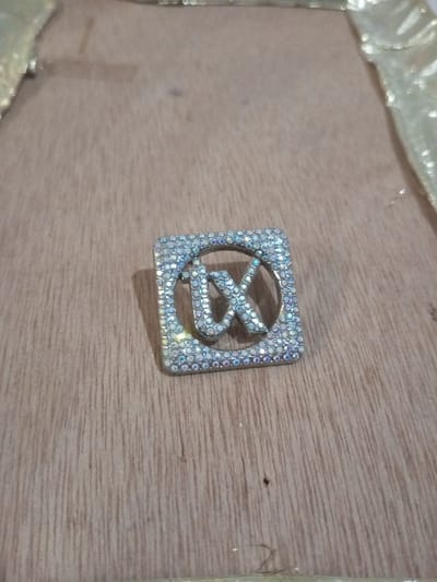 A small, square-shaped accessory is depicted lying flat on a light brown wooden surface with distinct vertical graining, in Chandragiri, Nepal. The accessory is silver-toned and intricately encrusted with numerous small, glittering, iridescent rhinestones. Its design features an outer square frame surrounding an inner stylized, lowercase "ux" inscription, also fully covered in rhinestones. Partial views of a crinkled, golden-colored fabric or ribbon are visible along the upper edges of the frame. No individuals are present, and no actions or interactions are occurring within the scene.