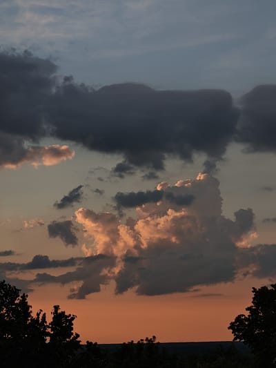 The image depicts a dramatic sunset over a rural landscape, likely in the vicinity of Семёновка, Russia. The sky is filled with a striking contrast of dark, brooding storm clouds and luminous, peach-colored cumulus clouds illuminated by the setting sun. The lower portion of the sky is a vibrant orange, transitioning to a pale blue-grey towards the top. Silhouetted against this colorful backdrop are the dark, jagged outlines of trees and a distant, dark horizontal line suggesting a horizon or a densely wooded area. There are no people, animals, or visible man-made structures in the image, emphasizing a natural and somewhat solitary scene. The overall impression is one of atmospheric intensity and the raw beauty of nature.