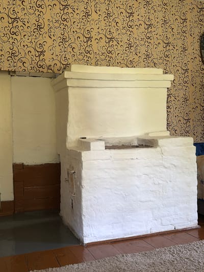 The image depicts an interior scene, likely a room in a house in Teterino, Russia, characterized by a traditional white-plastered stove that dominates the foreground. The stove, which appears to be a prominent feature of the dwelling, has a multi-layered structure with a large rectangular opening at the top, suggestive of a cooking or heating apparatus. It is constructed of rough, textured material and shows some signs of age and wear, with visible cracks in the plaster.

The walls of the room are covered in a distinctive wallpaper featuring a repeating pattern of ornate, brown swirls against a yellow background. To the left of the stove, a section of the wall is painted white, and below that, there is a wooden paneling painted a dark brown. The floor is a combination of polished wooden planks and a light-colored, textured rug.

The lighting in the room appears to be natural, casting soft shadows and highlighting the textures of the stove and wallpaper. There are no people or animals visible in the image, and no actions are being depicted. The scene conveys a sense of a lived-in, possibly older, home, with the stove serving as a focal point of the domestic environment. The overall impression is one of rustic simplicity and traditional Russian interior design.