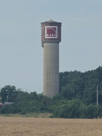 The image shows a tall, concrete water tower with a brick top section. A large, rectangular sign with a red background and white lettering that reads "AJAX" is visible on the upper portion of the tower. The tower is situated in a rural landscape with a field of dry crops in the foreground and a dense line of green trees in the background. A utility pole with wires runs along the right side of the image, near the trees. The sky is a pale, overcast blue. The water tower is located in Bozduganovo, Bulgaria.