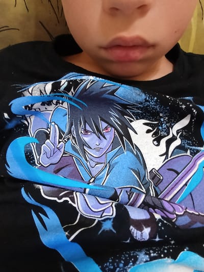 A close-up, top-down shot shows a child wearing a black t-shirt featuring a vibrant anime-style graphic. The graphic depicts a character with spiky dark hair, purple eyes, and light purple skin, dressed in blue and purple attire. The character is shown with a hand raised, making a gesture, and is surrounded by swirling blue energy and dark, starry background elements. The child's chin and mouth are visible in the upper part of the image, with their face cropped from view. The image does not depict any identifiable landmarks or specific environmental context that would allow for the determination of a location within Novotitarovskaya, Russia.