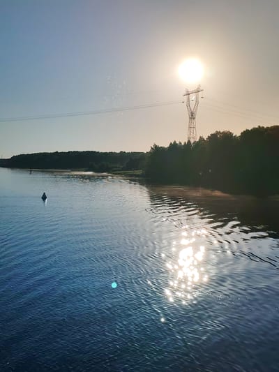 The image depicts a sunlit outdoor scene featuring a wide body of water, likely a river, with a shoreline bordered by trees. The location is specified as Gari, Russia.
In the upper right quadrant, the sun appears as an intense, circular white orb, casting a powerful glare. It is positioned directly behind the upper structure of a tall, lattice-style metal power transmission tower (pylon), silhouetting part of the tower. Multiple parallel power transmission lines extend horizontally across the upper middle portion of the sky, originating from or passing near the tower. The sky itself transitions from a clear, light blue on the left to a luminous, pale yellow-white near the sun. Several small, irregular white lens flares or atmospheric particles are visible against the sky, particularly above the power lines on the left.
A dense line of dark green deciduous trees forms the distant shoreline, stretching across the middle ground from the right towards the left. Between the water and the tree line, in the mid-left, a lighter-colored bank, possibly sandy or muddy, is visible.
The water in the foreground and mid-ground is a deep blue-teal, characterized by numerous small ripples on its surface, suggesting either a gentle current or a light breeze. A prominent, elongated patch of shimmering, sparkling sunlight reflects intensely off the water's surface, extending from the lower right foreground towards the center, directly beneath the sun's position. A solitary, conical white buoy floats in the mid-left section of the water, casting a subtle shadow. A distinct, bright turquoise/cyan circular light flare or artifact is present in the lower-middle left section of the water.
The scene is devoid of people, animals, or active human interactions, presenting a tranquil, natural environment juxtaposed with elements of industrial infrastructure.