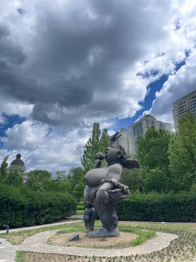 The image shows a bronze statue of a man carrying a lamb, located in Changping District, China. The statue stands on a small grassy area bordered by a curved stone path. In the background are tall trees and a building with a dome and ornate details, and a modern high-rise building with multiple windows. The sky is filled with clouds, with patches of blue visible. The statue appears to be weathered. The shot is taken from a low angle, looking up at the statue and the sky.