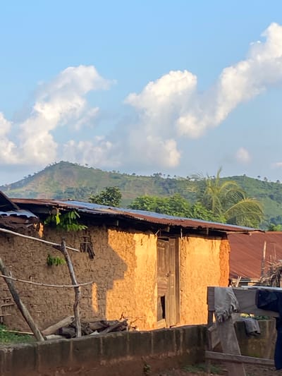 The image displays a rural scene in Igbara, Nigeria, under a clear blue sky with scattered white clouds. The perspective is angled, making it appear as if the horizon is tilted.

In the foreground, part of a building constructed with mud and rough textures is visible. The building has a rusty, corrugated metal roof. Adjacent to it, another structure with a reddish-brown roof and textured yellow walls is seen. Wooden planks form part of the building's facade, and there are dark openings suggesting windows or doorways. Some branches and vegetation are growing around and on the structures, indicating a natural integration with the environment.

In the background, a lush green hillside rises, dotted with trees and smaller vegetation. The terrain suggests a hilly or mountainous region. The sunlight casts shadows, indicating it is likely daytime, possibly late afternoon given the warm tones on the building. The overall impression is one of a traditional, possibly rustic, settlement nestled within a natural landscape. No people are visible in the image, and there is no visible text.