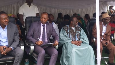 The image shows a group of men seated in chairs under a white canopy tent. The men are dressed in suits and traditional clothing. The men are all looking in different directions. There are several other people in the background, but they are not in focus. The setting is an outdoor event with green grass on the ground and white canopies. The location is likely in Rukoko, Democratic Republic of the Congo, based on the provided coordinates. The image suggests that the men are attending a formal event, such as a ceremony or a meeting. The man on the far left is wearing a blue shirt and a gray jacket. He is holding a black phone in his right hand and is sitting in a black chair. The man next to him is wearing a white shirt and a black suit. He is wearing a blue tie and holding a white pen in his right hand. The man is writing something on a white clipboard in front of him. The man next to him is wearing a green robe over a patterned shirt and brown vest. He has a white beard and mustache and is sitting with his legs crossed. The man next to him is wearing a brown suit and a white shirt. He is wearing a tan fedora hat and has glasses on. He is sitting with his legs crossed and holding a black phone in his right hand. The man is looking towards the front. The event appears to be held in an outdoor setting with green grass in the background. The image shows an event taking place in the Rukoko area of the Democratic Republic of Congo. The formal attire suggests an event of high importance. The men's serious expressions may indicate a business meeting, or a formal government event. The image could be a political rally, a ceremony, or a business meeting.