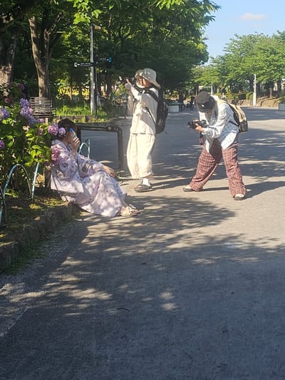 A person wearing a light-colored kimono with floral patterns sits on the ground next to a bench, holding a clear plastic bottle to their mouth. They have flowers in their hair. In the background, two other individuals are present. One person, wearing a light-colored wide-brimmed hat, a beige top, and pants, is holding a camera and appears to be taking a photo. They have a backpack on. Another person, wearing a plaid shirt, dark pants, and a black hat, is also holding a camera and looking at it. They also have a backpack. The scene appears to be a park in Taito, Japan, with trees and foliage providing shade. The ground is paved, and there are shadows cast by the trees.