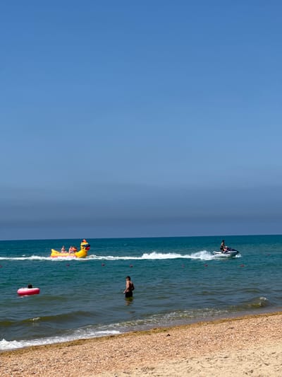 This media file captures a sunny day at the beach in Vityazevo, Russia. The scene is dominated by a vast expanse of clear blue sky meeting the turquoise ocean. The water is calm with gentle waves lapping at the shore, which is covered in fine, light-colored sand interspersed with pebbles and shells.

Several people are enjoying the water and beach. In the distance, a person is riding a jet ski, creating a spray of white water. Closer to the shore, a group of people are on an inflatable banana boat, which is yellow and shaped like a duck, with at least two people visible on it. Another person is seen in the water, standing waist-deep. Further out, another individual is swimming while holding onto a pink inflatable ring. A man in black swim trunks is standing in the shallow water near the beach.

The overall atmosphere is one of leisure and recreation. The bright sunlight suggests it is likely midday. The clear sky indicates fair weather. The presence of beachgoers, water sports, and inflatable toys all point to a typical summer day at a coastal resort. There is no visible text in the image.