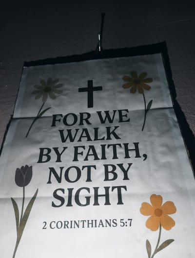 The image shows a close-up of a religious poster with text and floral illustrations. The poster is hung from a nail at the top. The text reads: "FOR WE WALK BY FAITH, NOT BY SIGHT" and below that "2 CORINTHIANS 5:7". There are drawings of flowers on either side of the text and a tulip on the left side. The poster appears to be made of paper and has some creases and discoloration. The background is a dark, textured wall. The lighting is dim, with shadows casting across the poster and wall. There are no people visible in the image. This image was taken in Iyafin, Nigeria.