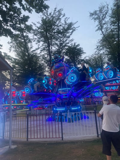 This media file captures a dynamic amusement park scene, likely in Manas, Kyrgyzstan, during the evening. The primary subject is a large, brightly lit amusement ride, dominated by vivid blue and red neon lights. The ride itself appears to be a spinning, multi-armed contraption with various pods or seats designed to hold passengers. Several people are visible, some riding the attraction and others standing nearby. One person in the foreground, wearing a white t-shirt and black pants, is looking towards the ride.
The setting is an outdoor amusement park, with trees visible in the background, their branches reaching towards a pale, twilight sky. The ground is covered in grass and a pathway or paved area, bordered by fences. The overall atmosphere suggests a lively evening at an amusement park, with the bright lights of the ride contrasting with the fading daylight. The mood appears to be one of excitement and enjoyment, typical of such an environment. There is no discernible text in the image, aside from a small logo or text on the white shirt of the person in the foreground, which is too blurry to read. The time of day is clearly evening, as indicated by the soft light and the illuminated nature of the ride.