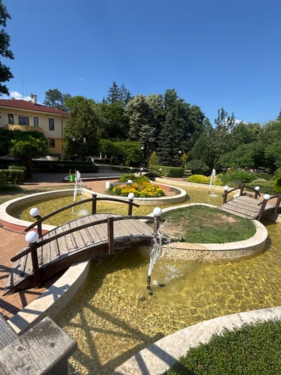 The image depicts a public park area in the city of Chavdar, Bulgaria, on a clear, sunny day. The foreground features a multi-tiered water feature with several shallow pools. Wooden bridges with white globe-shaped lights cross over these pools. Fountains are actively spraying water into the pools, creating ripples and splashing effects. The water itself appears a greenish-yellow hue. The edges of the pools are lined with light-colored tiles and some sections are adorned with mosaic tiles. Small islands within the pools are covered with grass.
To the left of the water feature is a paved walkway made of reddish-brown bricks. Beyond the walkway and to the left of the water feature is a light yellow building with a red-tiled roof and dark-framed windows. The building is partially obscured by lush green trees and bushes.
The background is dominated by a variety of trees, including evergreens and deciduous trees, forming a dense green canopy. The sky is a vibrant, cloudless blue. There are no people visible in the image. The overall scene conveys a sense of a well-maintained, tranquil park environment.