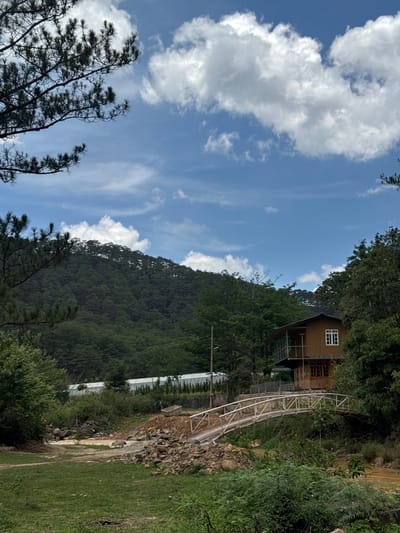 The scene is a rural area near Păng Tiêng 1, Vietnam. A wooden house with a white-framed window sits on a hillside, partially obscured by trees. A metal bridge leads from the house down the slope. Below the bridge is a rocky area with some construction. Beyond this, there are greenhouse-like structures. The sky is blue with scattered white clouds. In the foreground, there is grassy ground to the right and trees to the left.